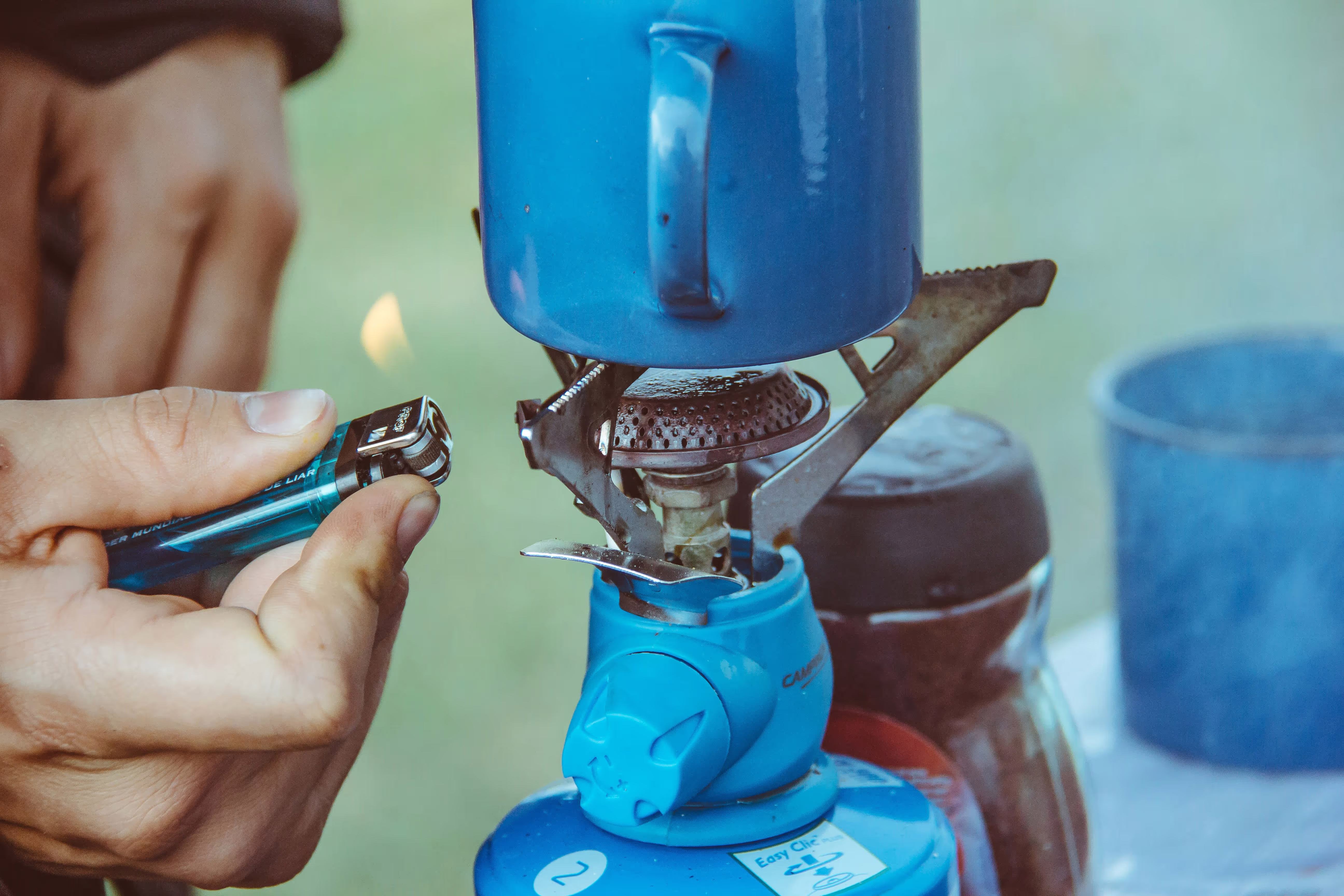Nahaufnahme einer Hand mit Feuerzeug, die einen Gaskocher anzündet, um eine Tasse Kaffee zuzubereiten.